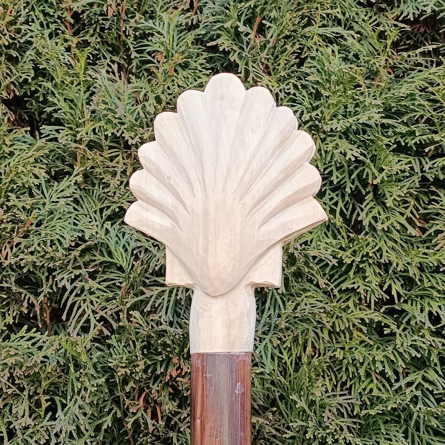 Kopf eines Pilgerstabs, mit Jakobsmuschel aus hellem Holz, vor gr&uuml;ner Tujahecke
