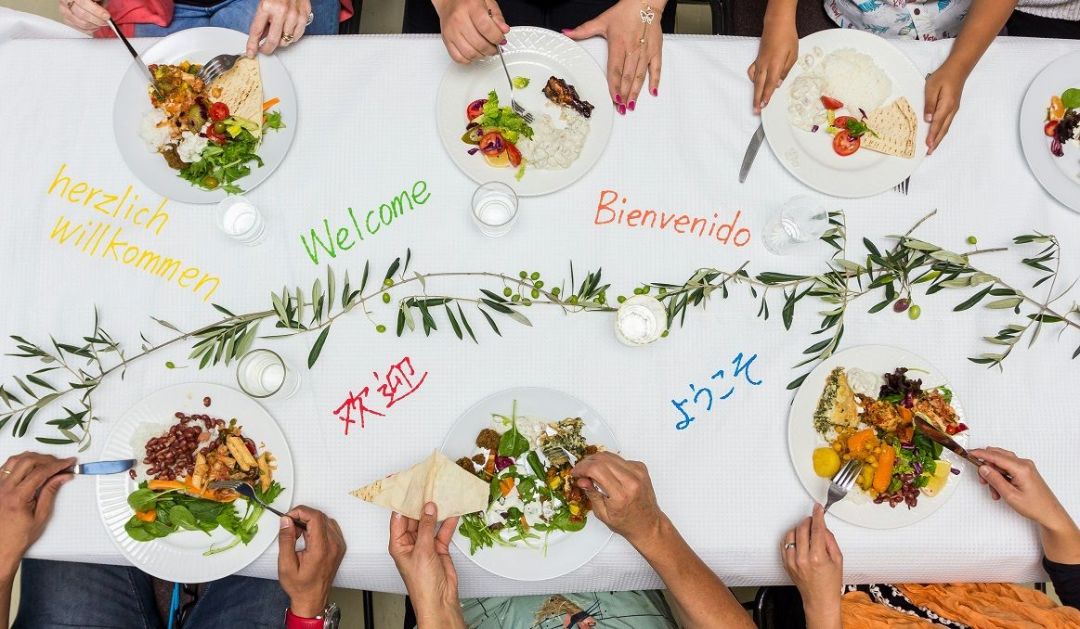 Menschen sitzen um einen Tisch. Vor Ihnen stehen Teller mit Essen. Auf dem Tischtuch steht Willkommen in unterschiedlichen Sprachen.